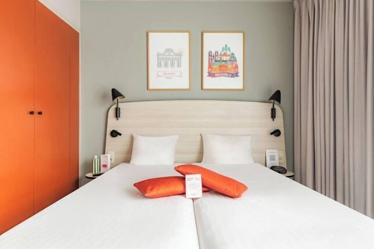 Ein Hotelzimmer mit einem Bett, zwei gerahmten Bildern an der Wand, einem Nachttisch und einem orangefarbenen Kleiderschrank.