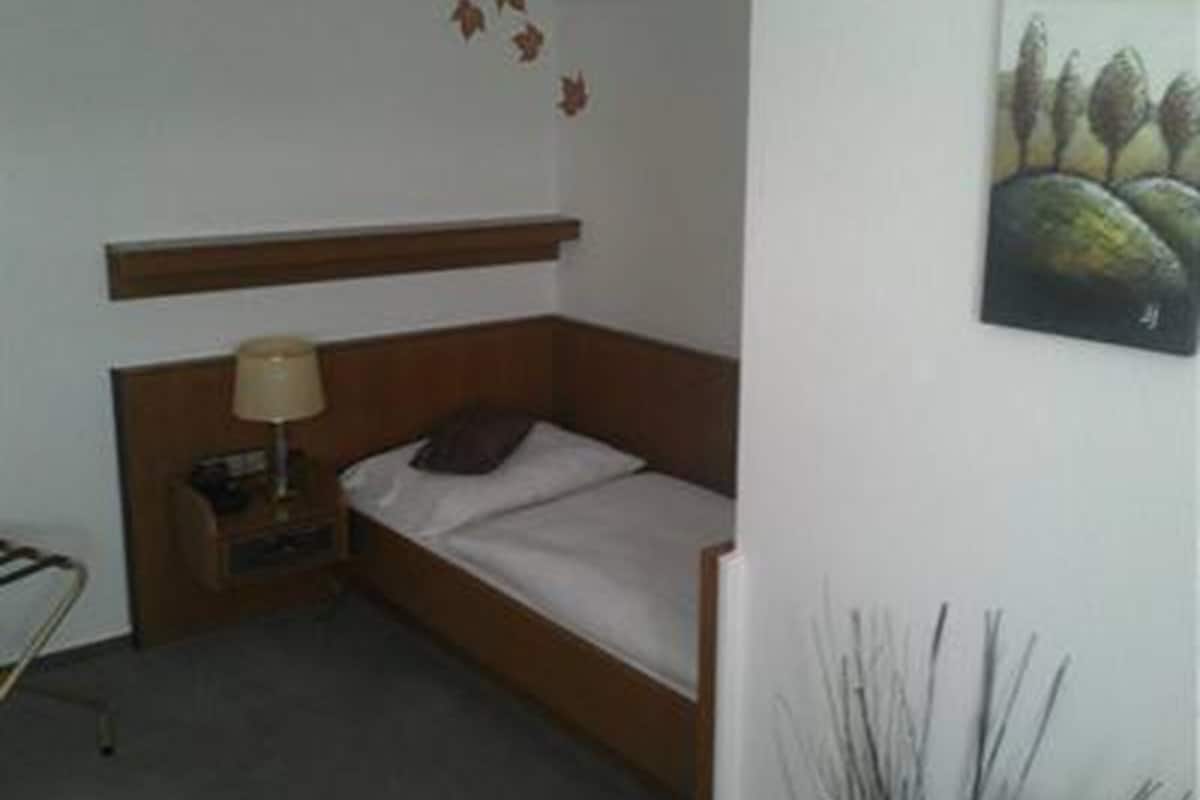 Un lit simple avec une tête de lit en bois, une table de chevet avec une lampe et une œuvre d’art fixée au mur représentant des arbres.