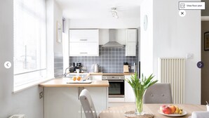 A modern kitchen with white cabinets, a built-in oven, and a coffee machine. There is a dining table with a vase of flowers and a plate of fruit. - Putney Court - 1 Bed Apartment by BaseToGo (London)