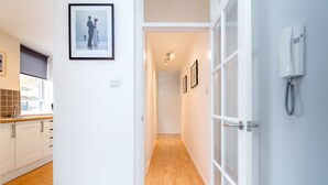 A hallway with a wooden floor leading to a kitchen area with white cabinets and a window. - Putney Court - 1 Bed Apartment by BaseToGo (London)