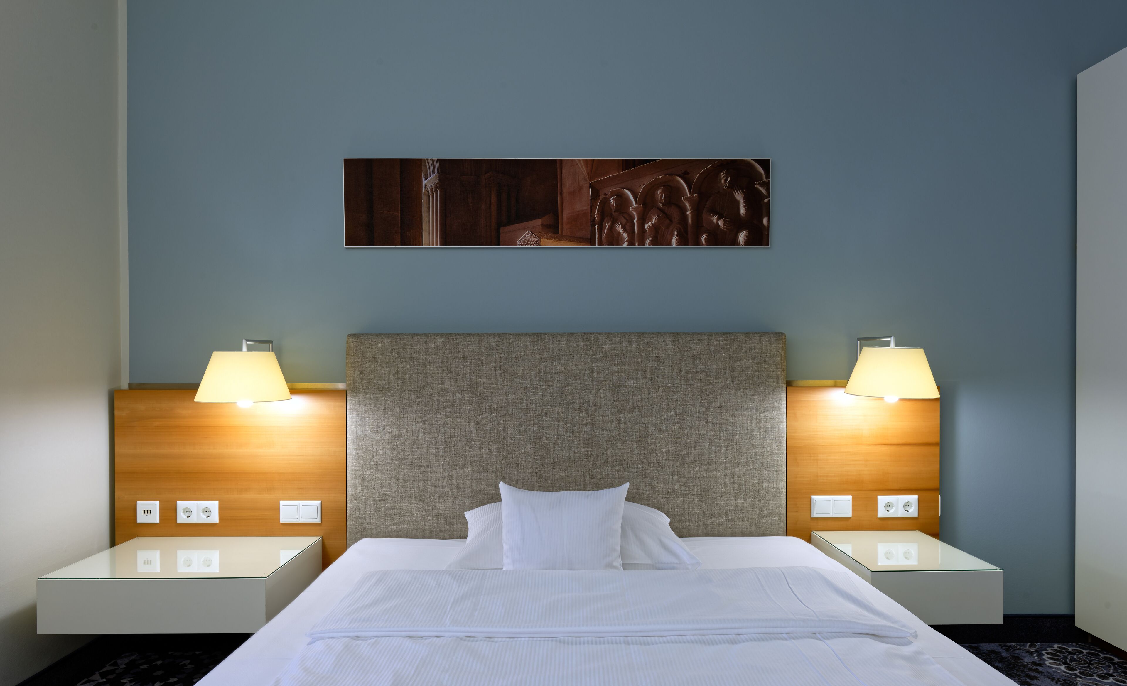 Habitación de hotel con una cama grande, dos lámparas de noche, una obra de arte en la pared y un cabecero de madera.