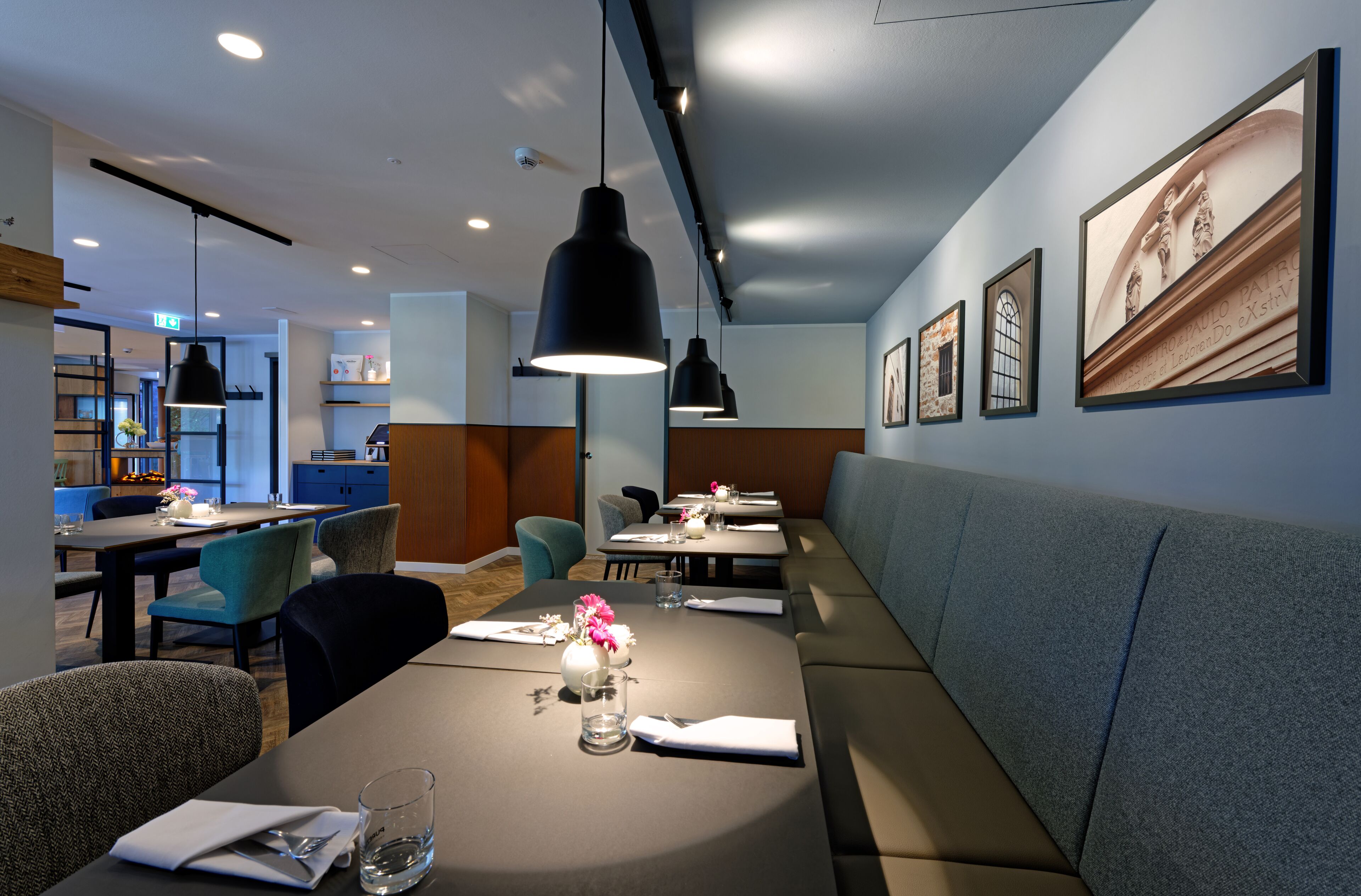 Ein modernes Restaurant mit hohen Tischen, gemütlicher Sitzgelegenheiten, Pendelleuchten und gerahmten Kunstwerken an den Wänden.