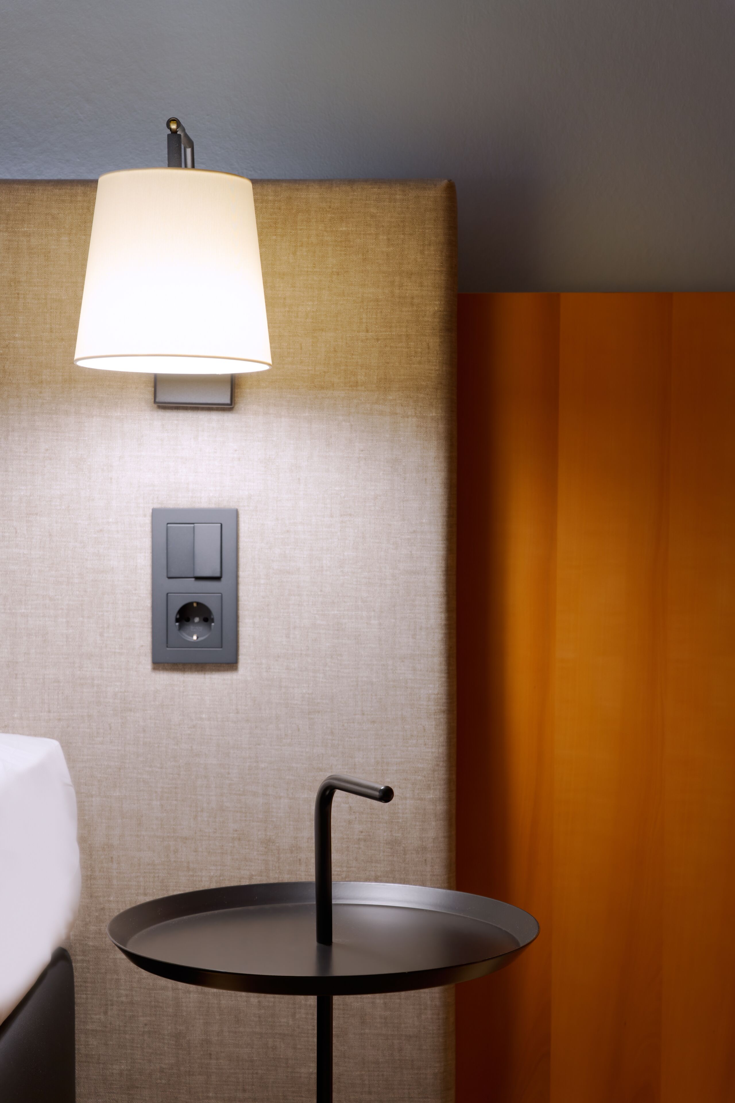 A hotel room with a round table, a wall-mounted lamp, and a power outlet.