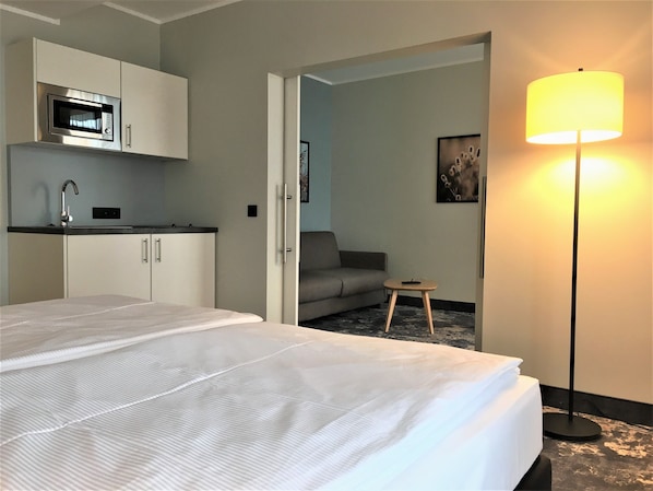 A hotel room with a bed, a kitchenette with a microwave, a sofa, a small table, and a floor lamp. - Hotel Am Kloster (Werne)