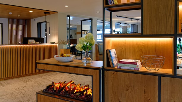 A modern reception area with a wooden reception desk, a fire feature, and a bookshelf with books and decorative items. - Hotel Am Kloster (Werne)