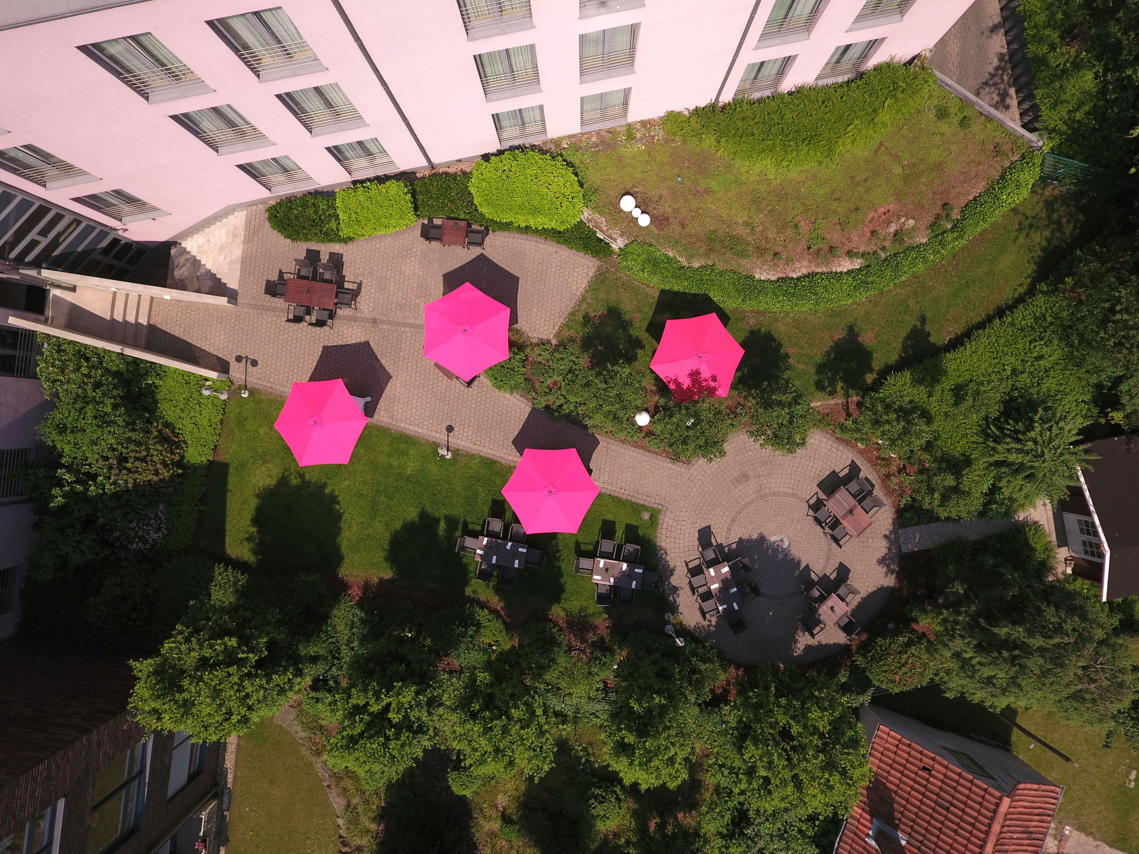 Vista aérea de un patio con mesas hexagonales y sombrillas rosas, rodeado de vegetación y edificios.