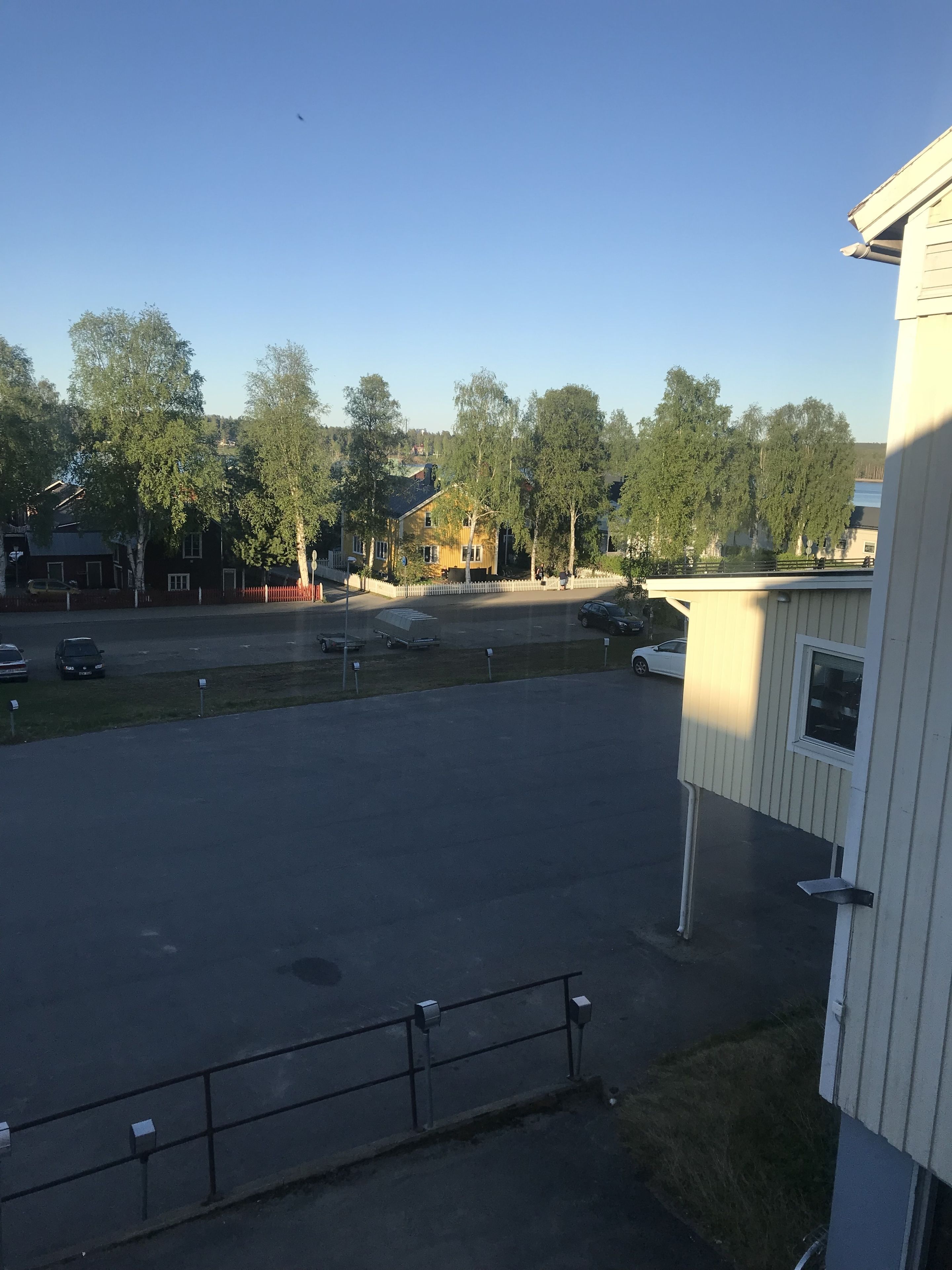 A parking lot with several cars, a white building with a window, and trees in the background.