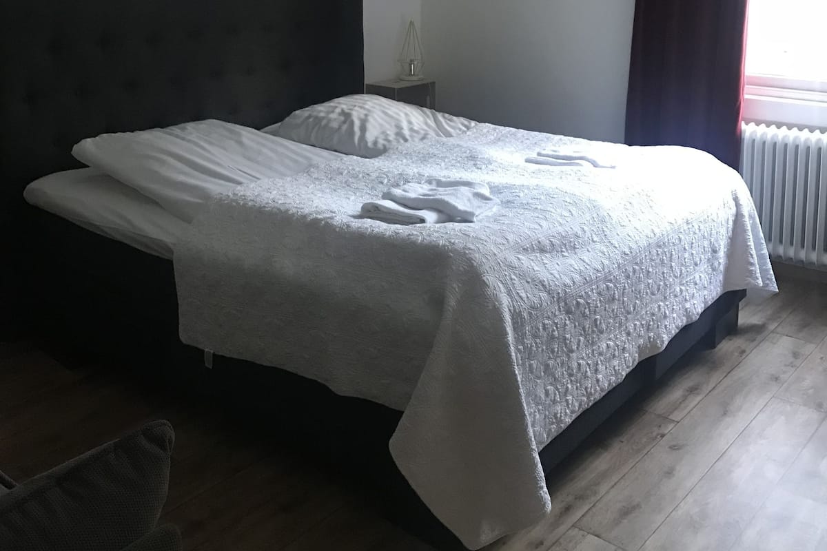 A bedroom with a large bed, a wall clock, a radiator, and a window with curtains.