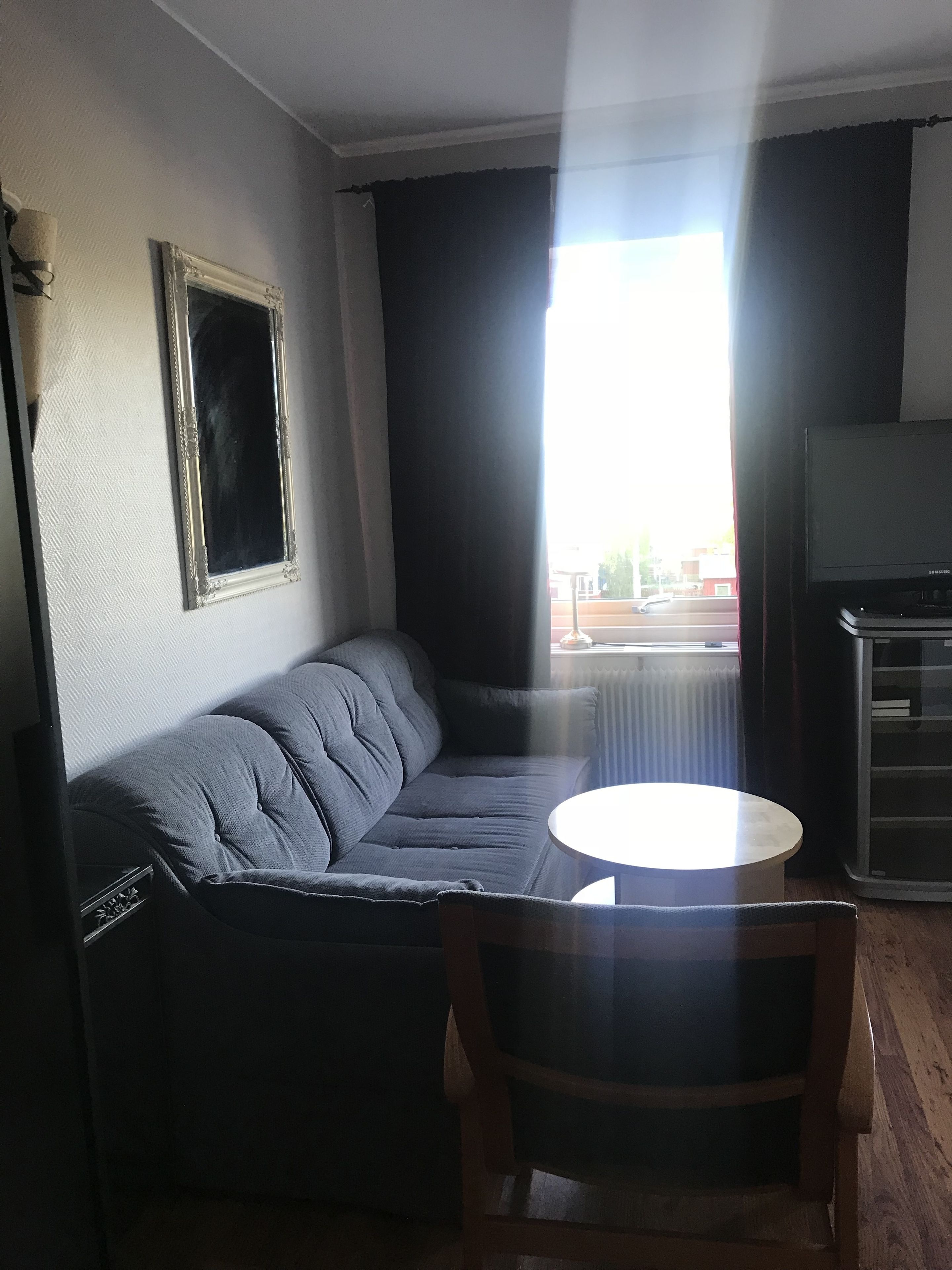 Een woonkamer met een grijze bank, een ronde tafel, een houten stoel, een televisie en een raam met gordijnen.