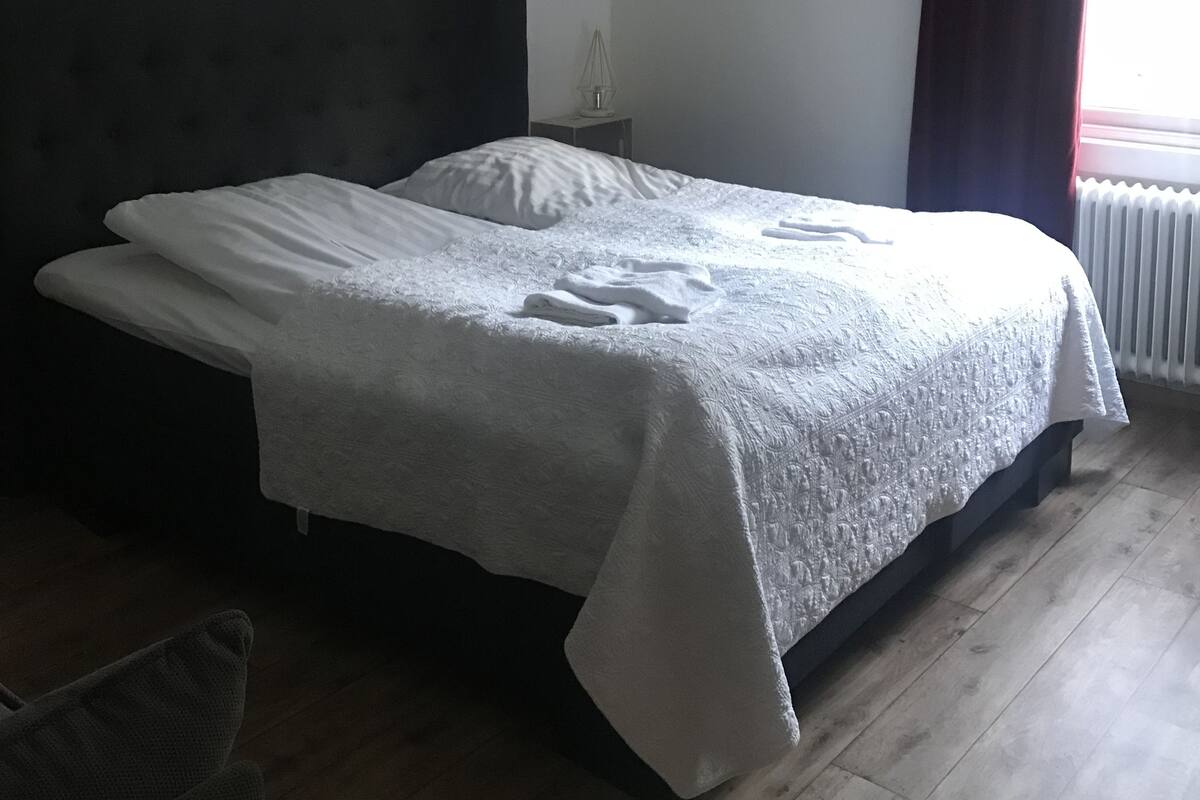 Ein Schlafzimmer mit einem großen Bett, einer Wanduhr, einer Heizung und einem Fenster mit Vorhängen.