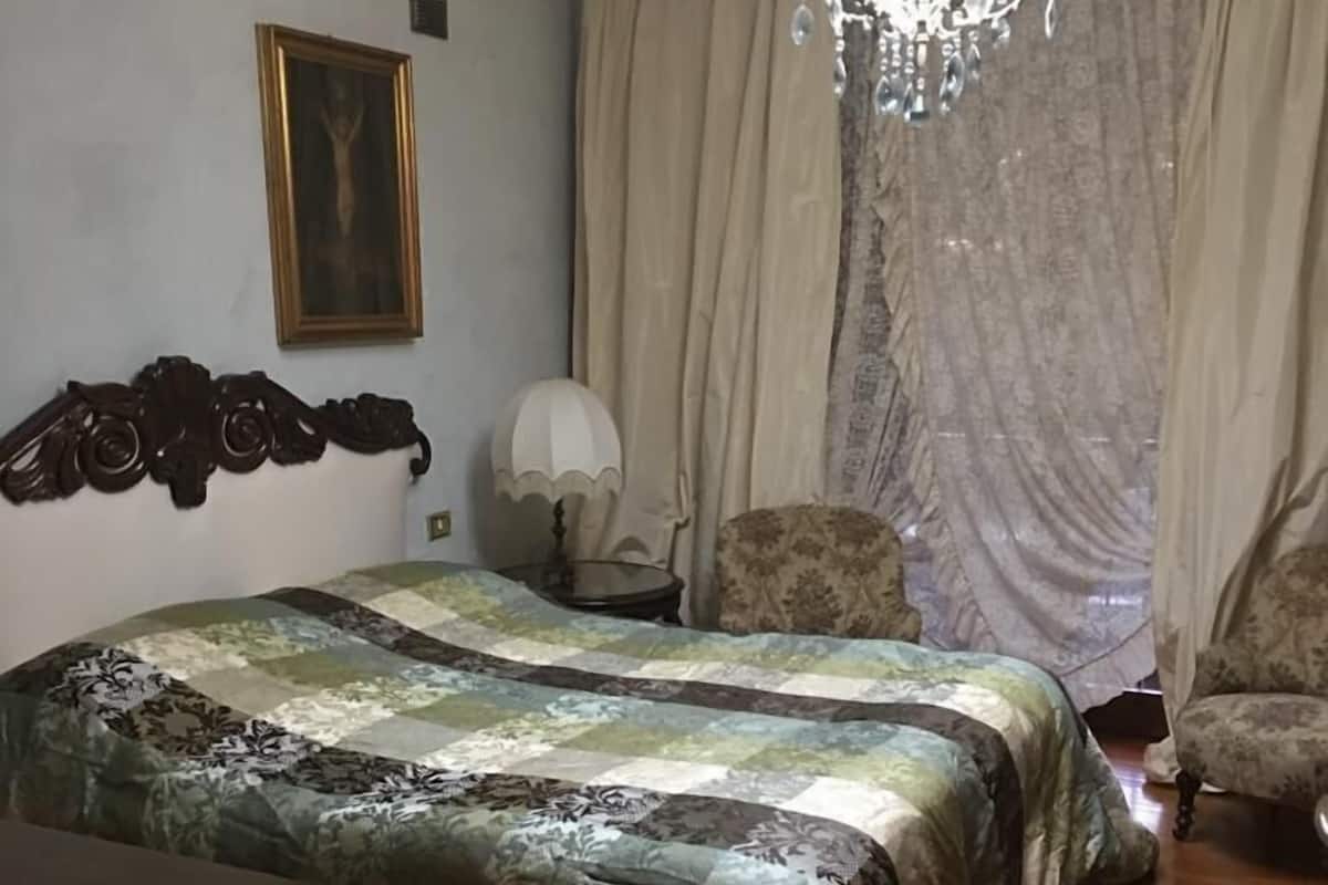 A bedroom with a bed, a chair, a chandelier, curtains, and a framed picture on the wall.