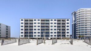 A modern multi-story hotel with balconies, located on a sandy beach with a clear sky.