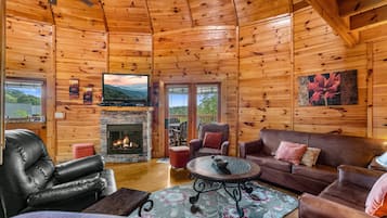L’intérieur chaleureux d’un chalet avec une cheminée, des fauteuils en cuir, un téléviseur à écran plat et une grande fenêtre donnant sur un paysage verdoyant.