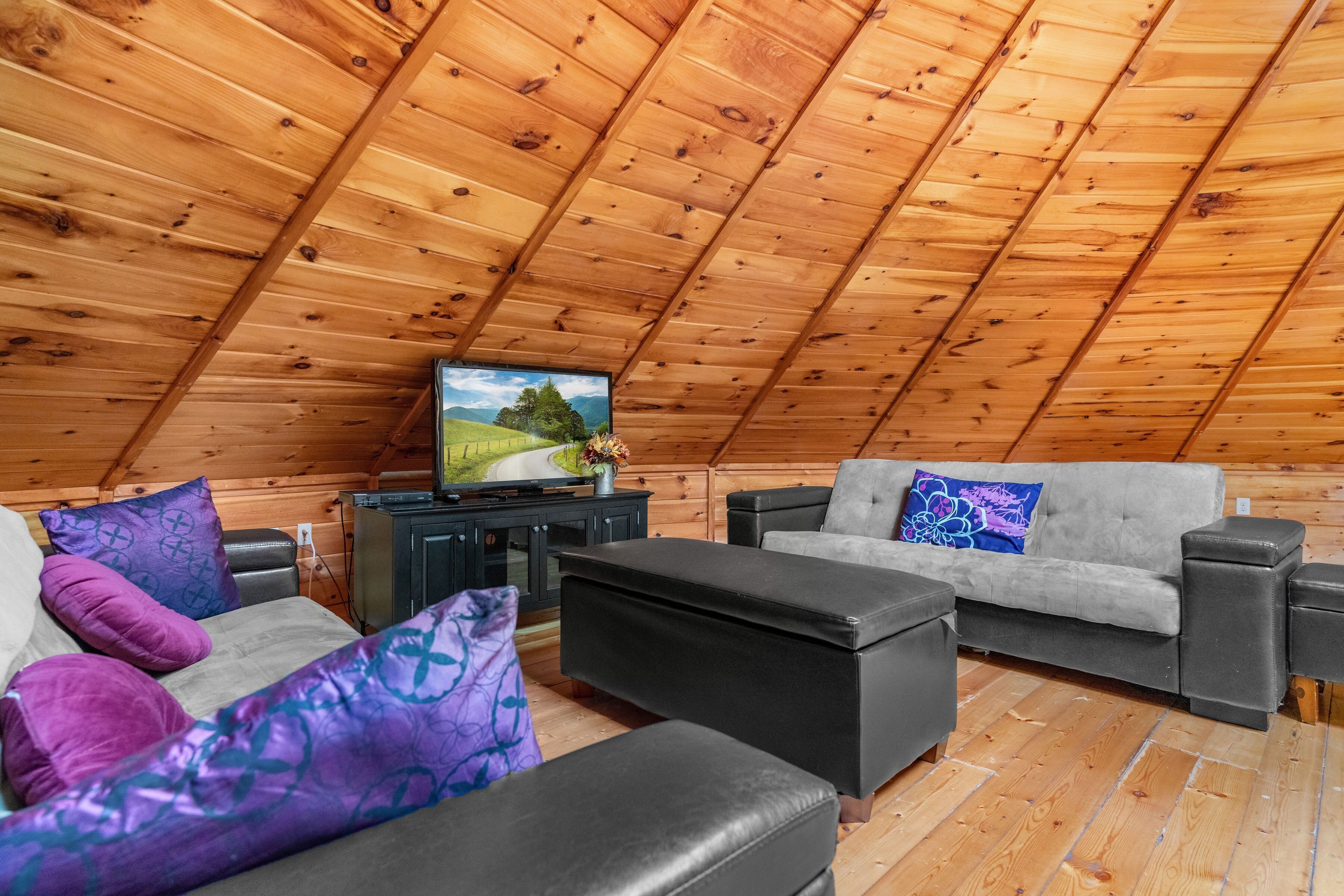 A cozy attic room with a wooden sloped ceiling, a television on a wooden stand, a grey sofa, and a black ottoman.