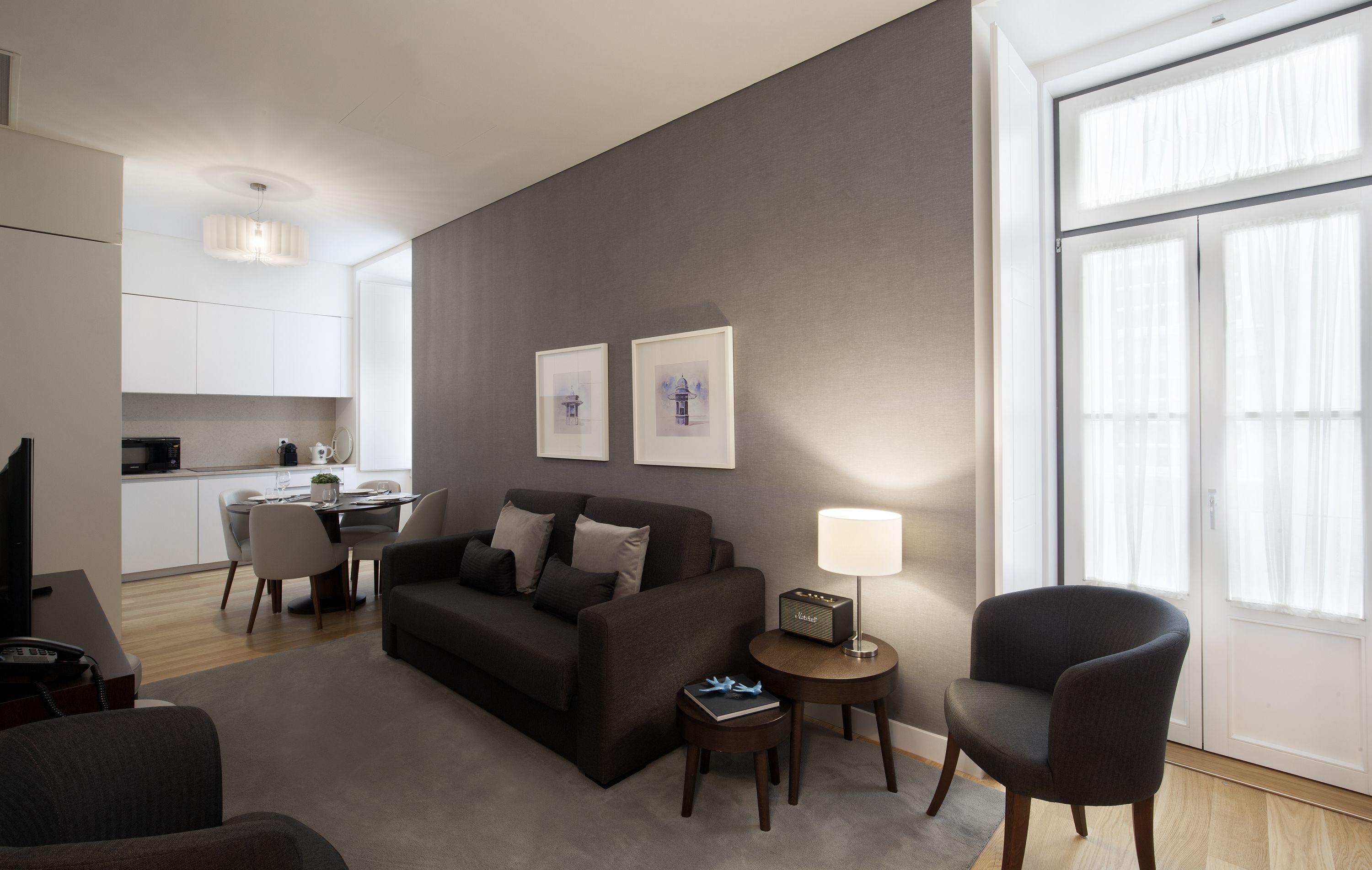 Una sala de estar moderna con un sofá, una mesa de centro, una mesita de noche con una lámpara y un sillón. Hay una zona de comedor con una mesa y sillas, y una cocina al fondo.