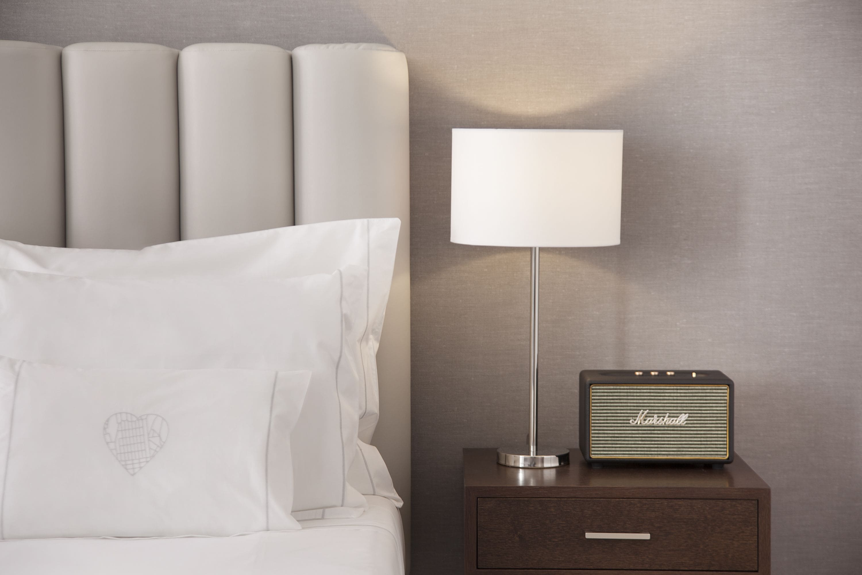A neatly made bed with white linens and pillows, a bedside lamp, and a Marshall radio on a wooden nightstand.