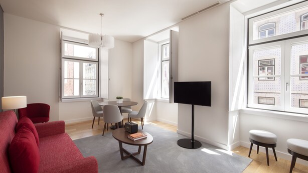 Una sala de estar moderna con un sofá rojo, una mesa de centro redonda y un comedor con mesa y sillas. Hay un televisor de pantalla plana montado en la pared, y ventanas grandes que permiten la entrada de luz natural.
