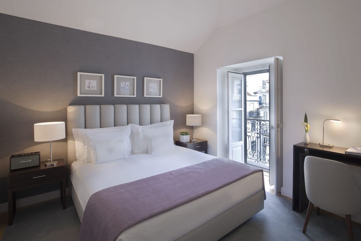 A hotel room with a bed, bedside lamps, a desk, a chair, and a balcony with a railing.