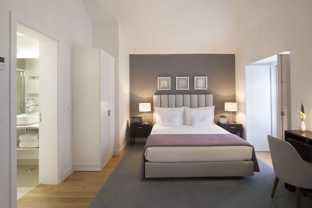 A hotel room with a bed, bedside tables, a desk, a chair, and a bathroom visible through an open door.
