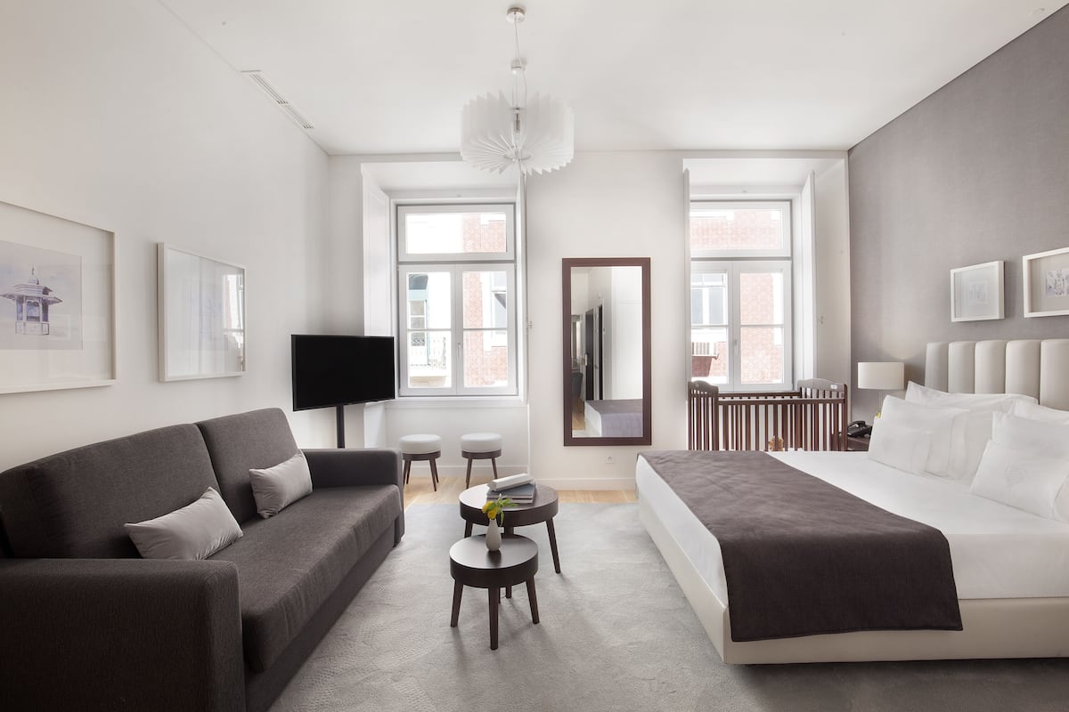 A modern hotel room with a grey sofa, a bed with white linens, a small round table with a vase, and a large mirror.