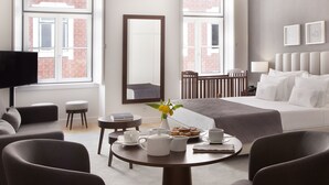 Una habitación de hotel moderna con una cama grande, un espacio de descanso con una mesa y sillas, un televisor y una ventana con vista a un edificio de ladrillo.