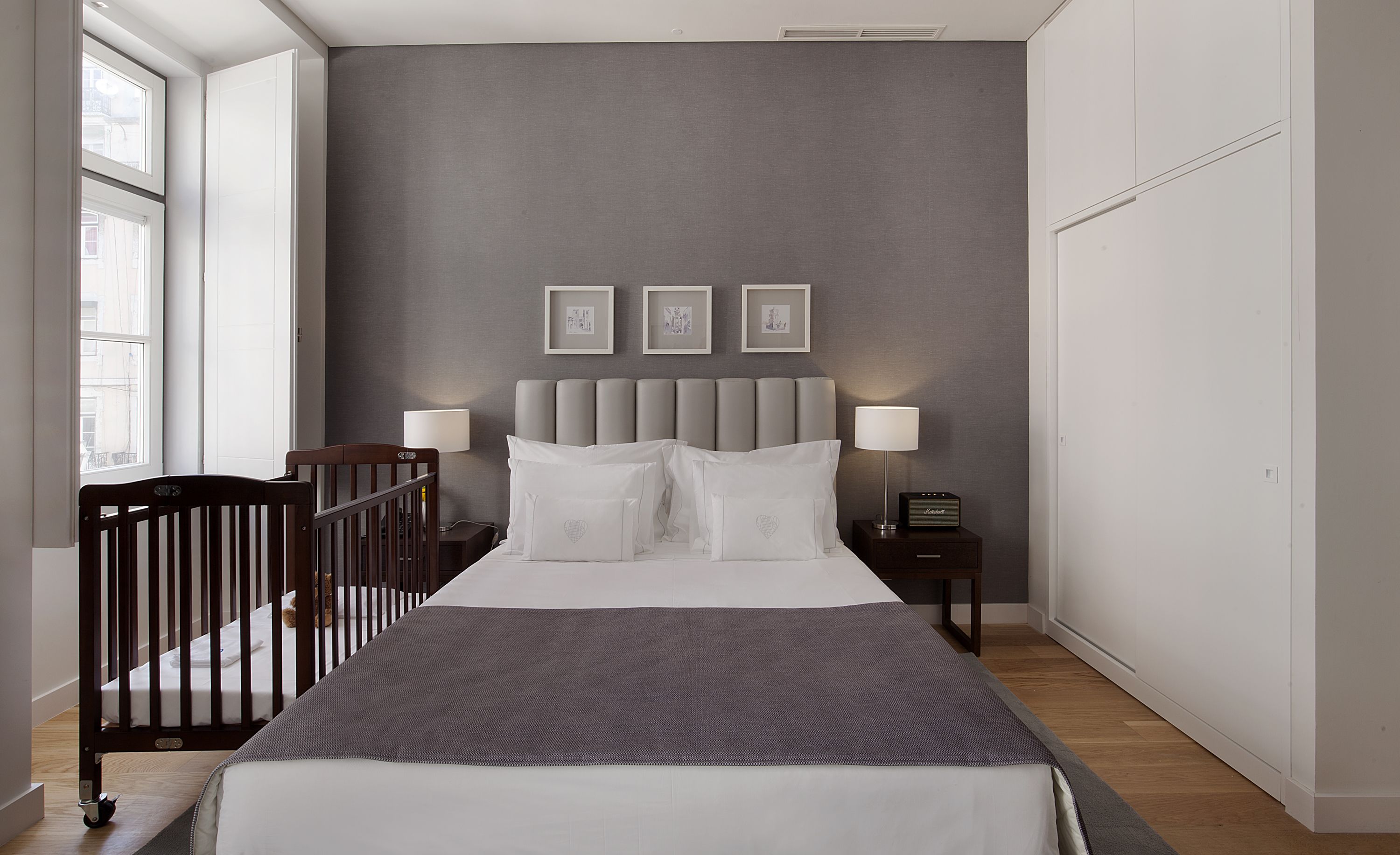 A bedroom with a bed, bedside lamps, a nightstand, a crib, and framed pictures on the wall.