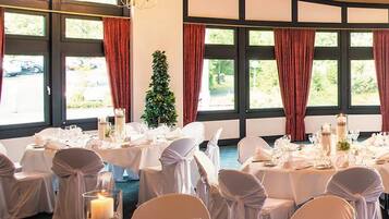 A banquet hall with round tables set for dining, large windows with curtains, and a chandelier.