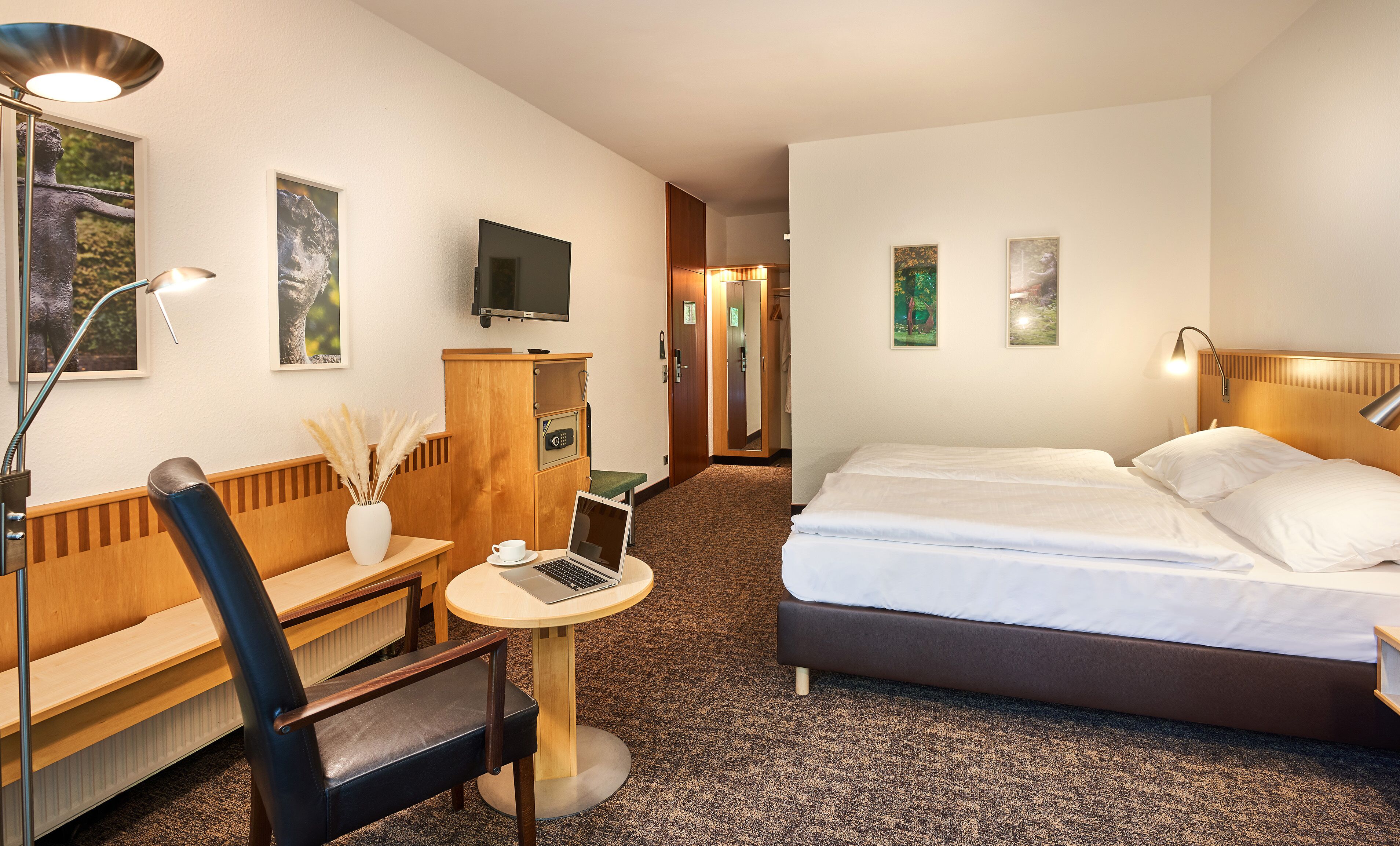 A hotel room with a bed, a desk with a chair, a small table with a cup and a laptop, a wooden bench, and a TV mounted on the wall.
