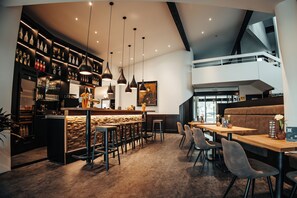 Un moderno bar con techo alto, vigas a la vista y una variedad de botellas y copas en estantes. Hay varias zonas de estar con mesas y sillas, y un gran ventanal que proporciona luz natural.