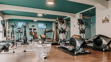 Une salle de sport équipée de divers appareils d’exercice, dotée d’un plancher en bois, de murs de couleur turquoise et d’un plafond avec des luminaires encastrés.