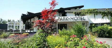 Un edificio moderno con un letrero que dice "PARK-HOTEL" y un jardín bien cuidado con diversas plantas y flores.