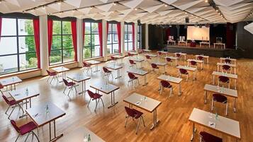Ein Konferenzraum mit mehreren Tischen und Stühlen, großen Fenstern mit roten Vorhängen und einer Bühne mit Projektor.
