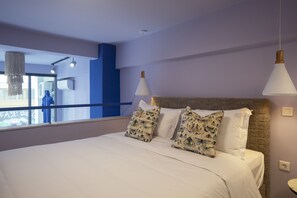 A modern bedroom with a large bed, two decorative pillows, a wicker headboard, and a view of a cityscape through a large window. - The Frogs Guesthouse (Athens)