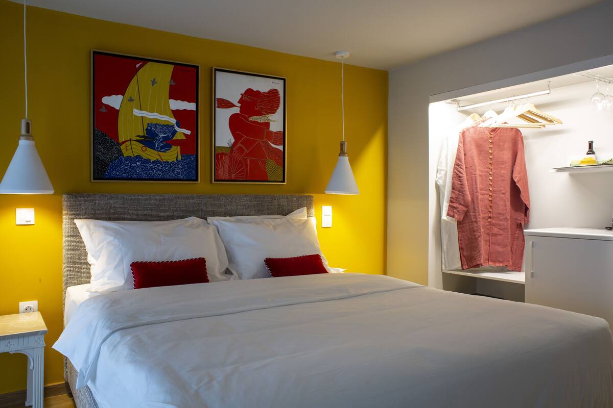 Quarto de hotel com uma cama, duas pinturas, um guarda-roupa e uma mesa de cabeceira.