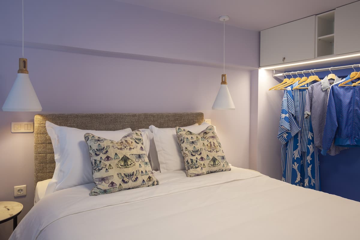 Cama bem arrumada com almofadas estampadas, mesa de cabeceira e um cabideiro com roupas penduradas.