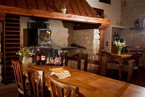 A rustic dining area with a wooden table, chairs, and a stone wall. There are wine bottles and glasses on the table, and a vase with flowers.