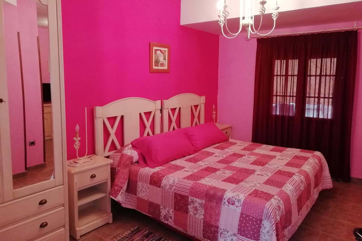 Une chambre à coucher comprenant un lit, une table de chevet, une commode, un miroir, un lustre et une fenêtre avec des rideaux.