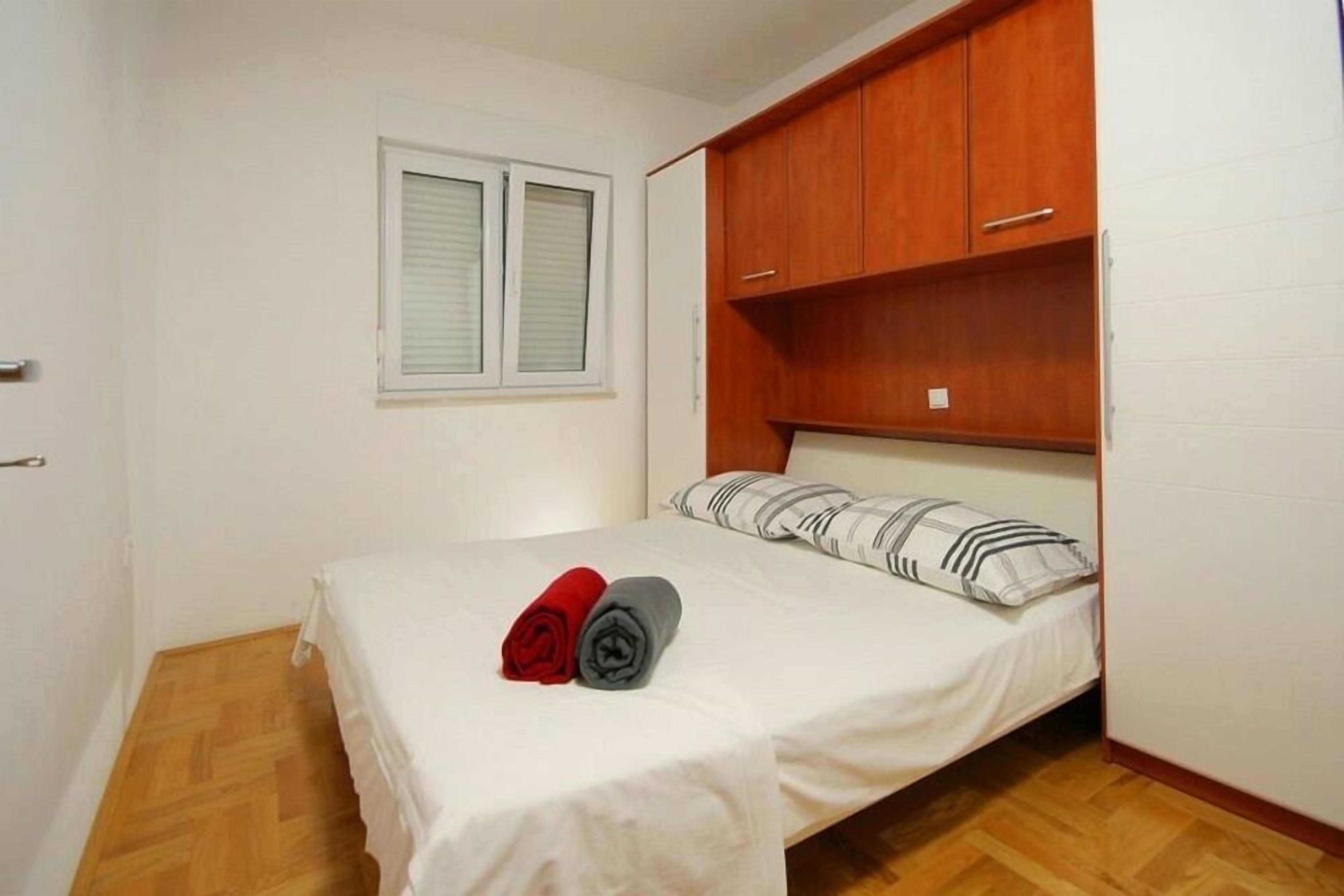 A single bed with a white mattress, two rolled towels (one red, one gray), and a striped pillow. There is a window with blinds, wooden cabinets above the bed, and a white wall.