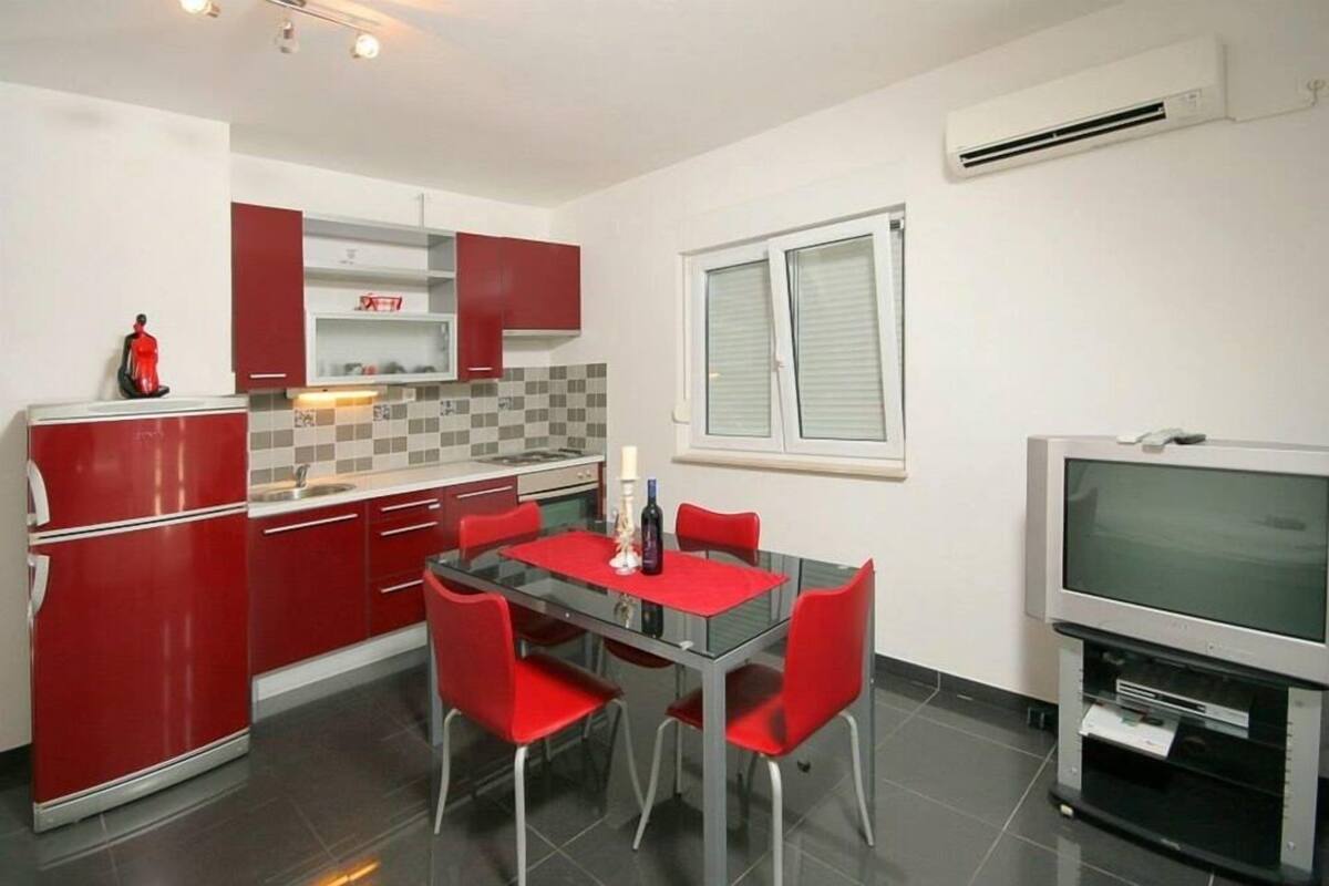 Una cocina compacta con armarios rojos, una mesa de comedor de vidrio y un mueble para televisión.