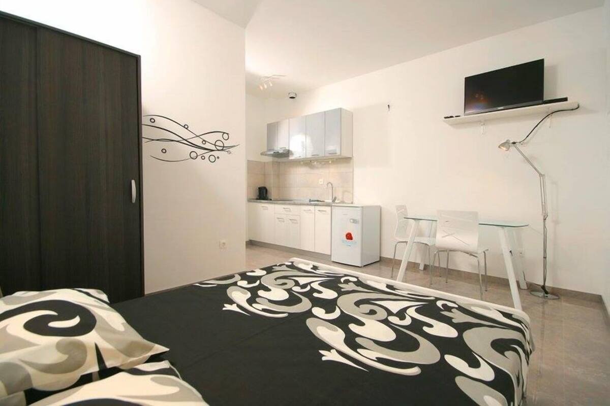 Una habitación compacta con cama, una kitchenette pequeña, un televisor montado en la pared y un escritorio con sillas.