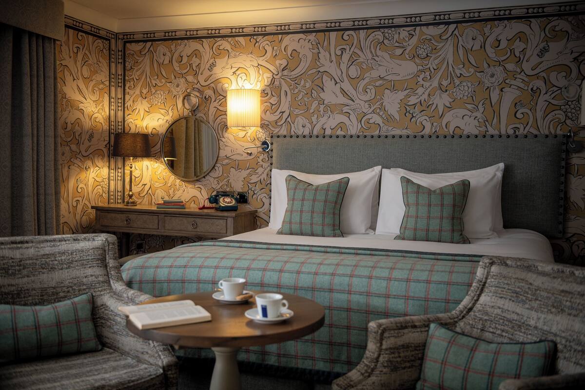 Habitación de hotel con una cama grande, dos almohadas, una mesita de noche con una lámpara, un espejo y una pequeña mesa redonda con dos tazas y un libro.