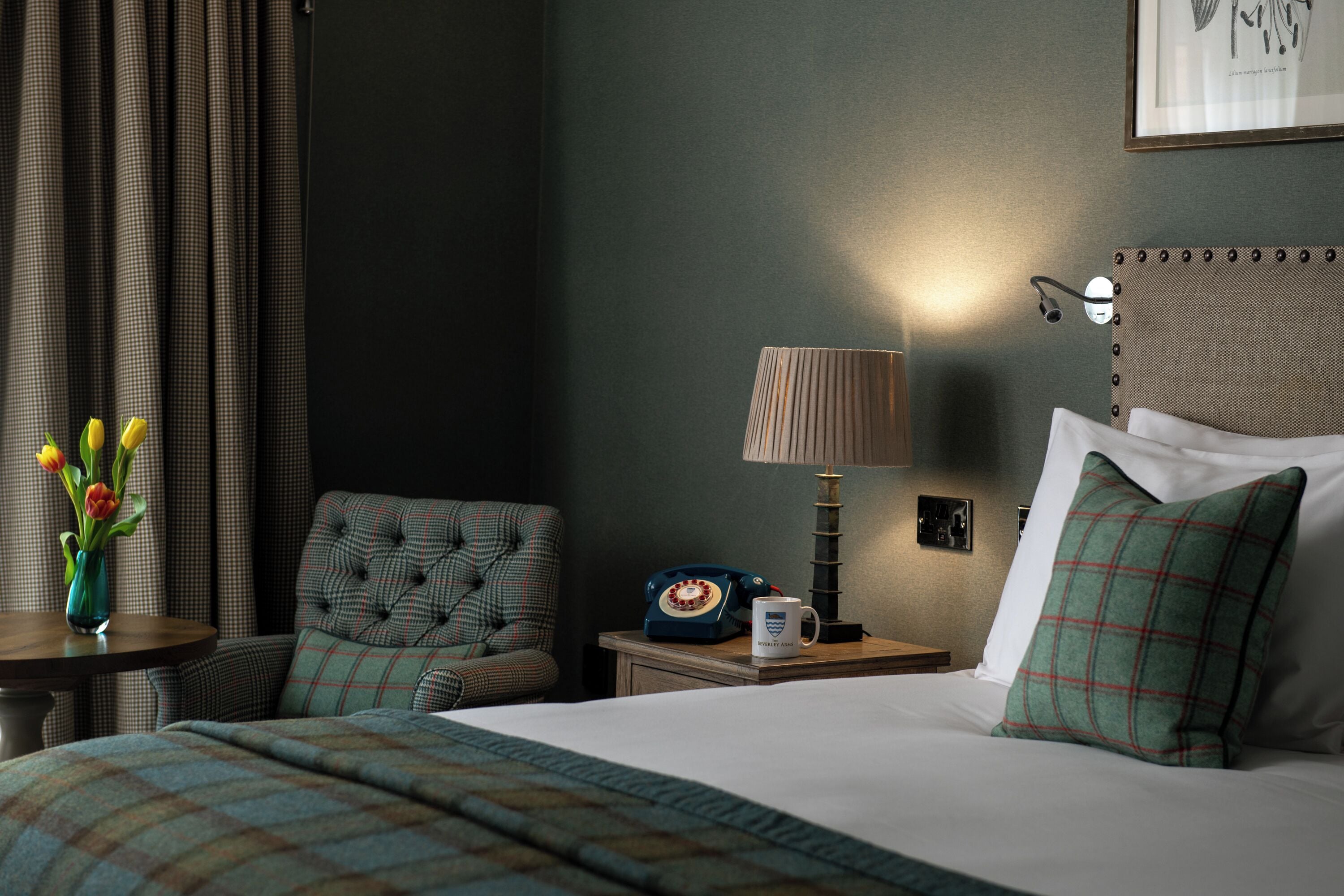 A hotel room with a bed, a chair, a table with a vase of flowers, a lamp, and a telephone.