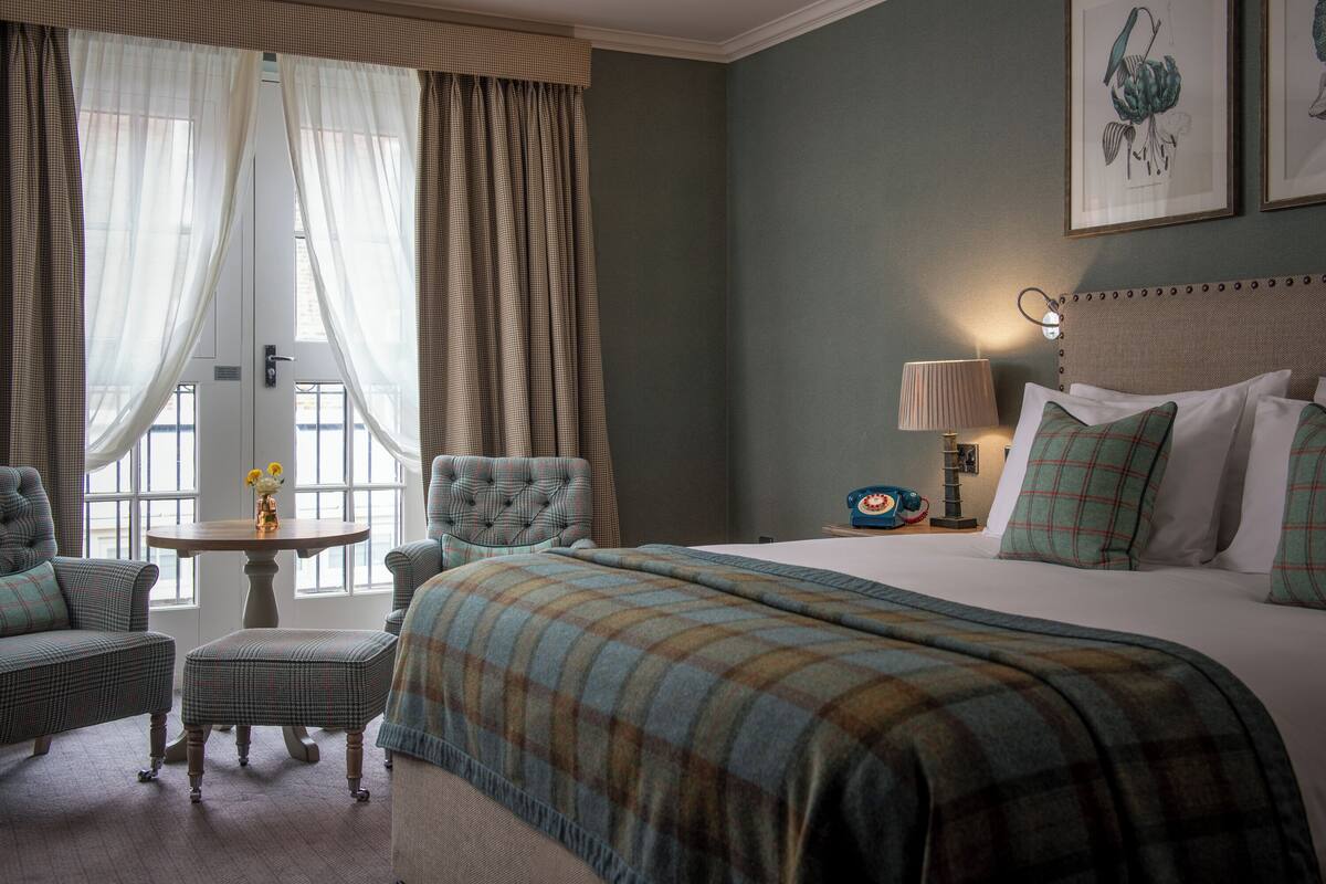 Habitación de hotel con una cama grande, dos sillones, una mesita con un florero y una lámpara en una mesita de noche.