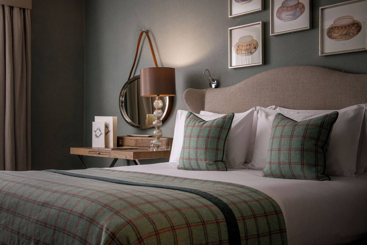 Una cama bien hecha con ropa de cama escocesa, una lámpara de noche y una mesita de noche con un cuadro enmarcado y un pequeño adorno.