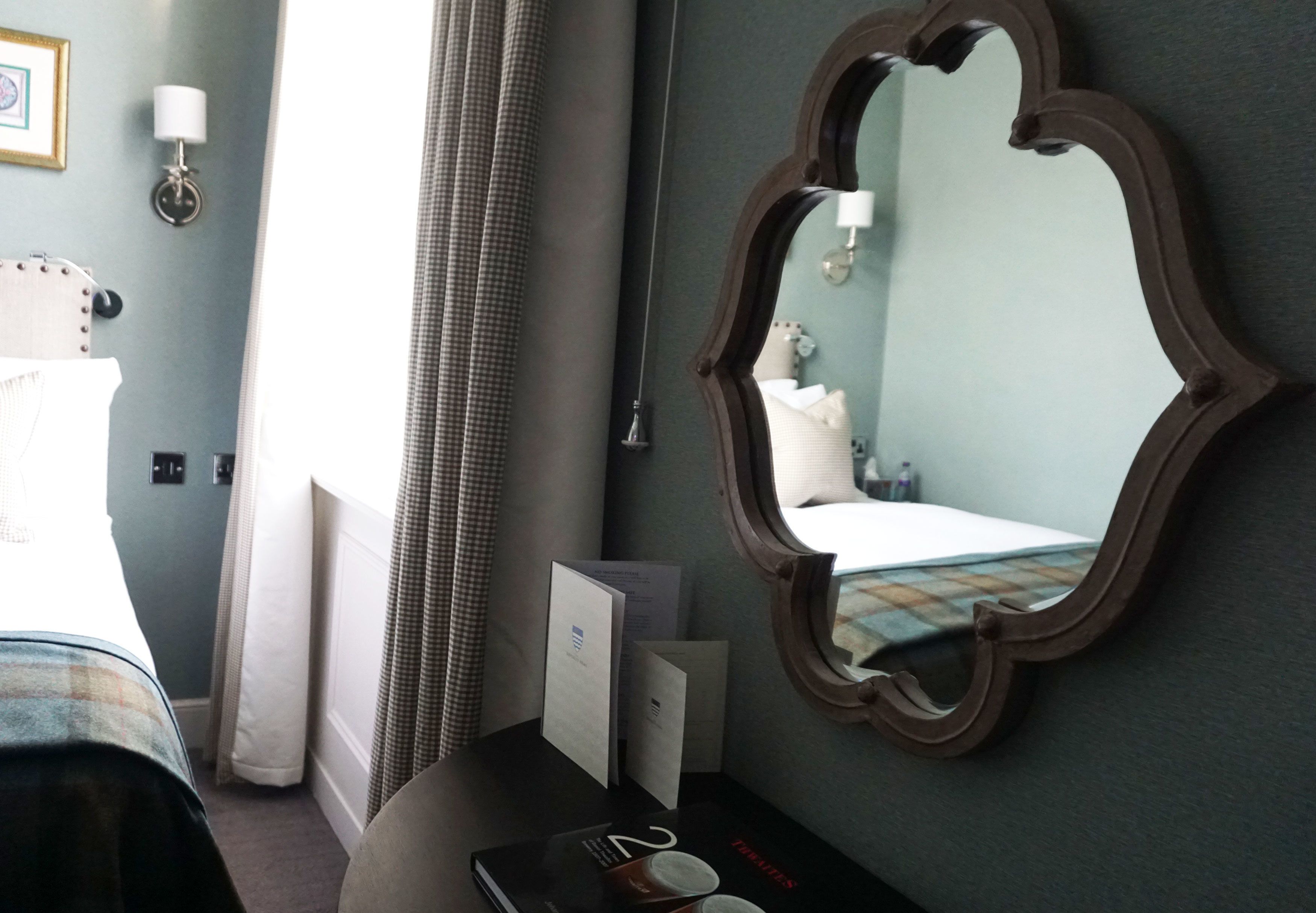 Habitación de hotel con una cama, una mesita de noche con lámpara, un espejo y una ventana con cortinas.