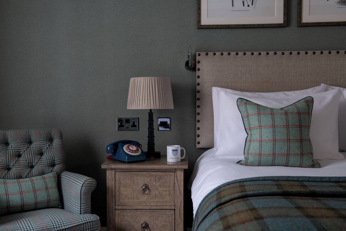 Habitación de hotel impecable con un sillón de estampado escocés, una mesita de noche con lámpara, un teléfono antiguo y una cama con manta y almohada, ambos de estampado de cuadros verdes.