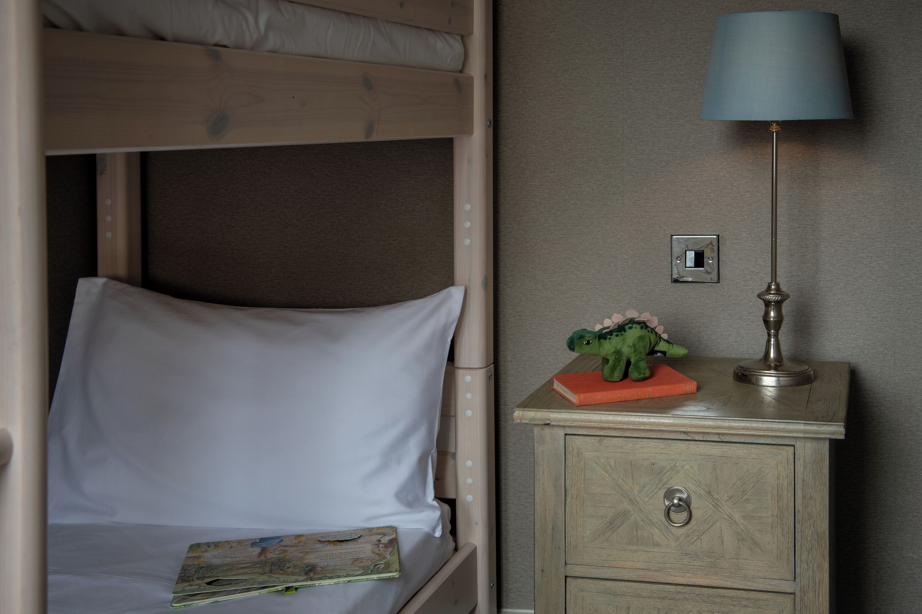Een stapelbed met een witte matras, een klein nachtkastje met een lamp, en een speelgoeddinosaurus op het tafeltje.