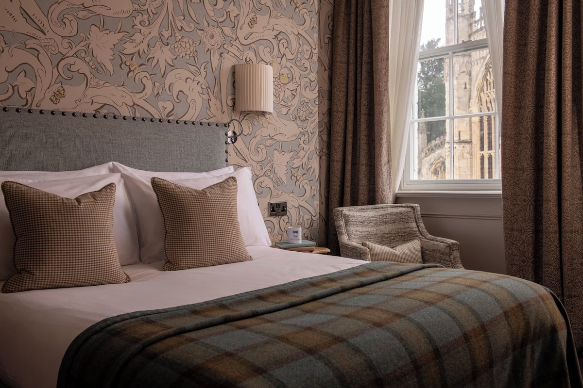 Habitación de hotel con una cama grande, almohadas a cuadros, una manta escocesa, una mesita de noche con lámpara y vista a un edificio a través de una ventana.