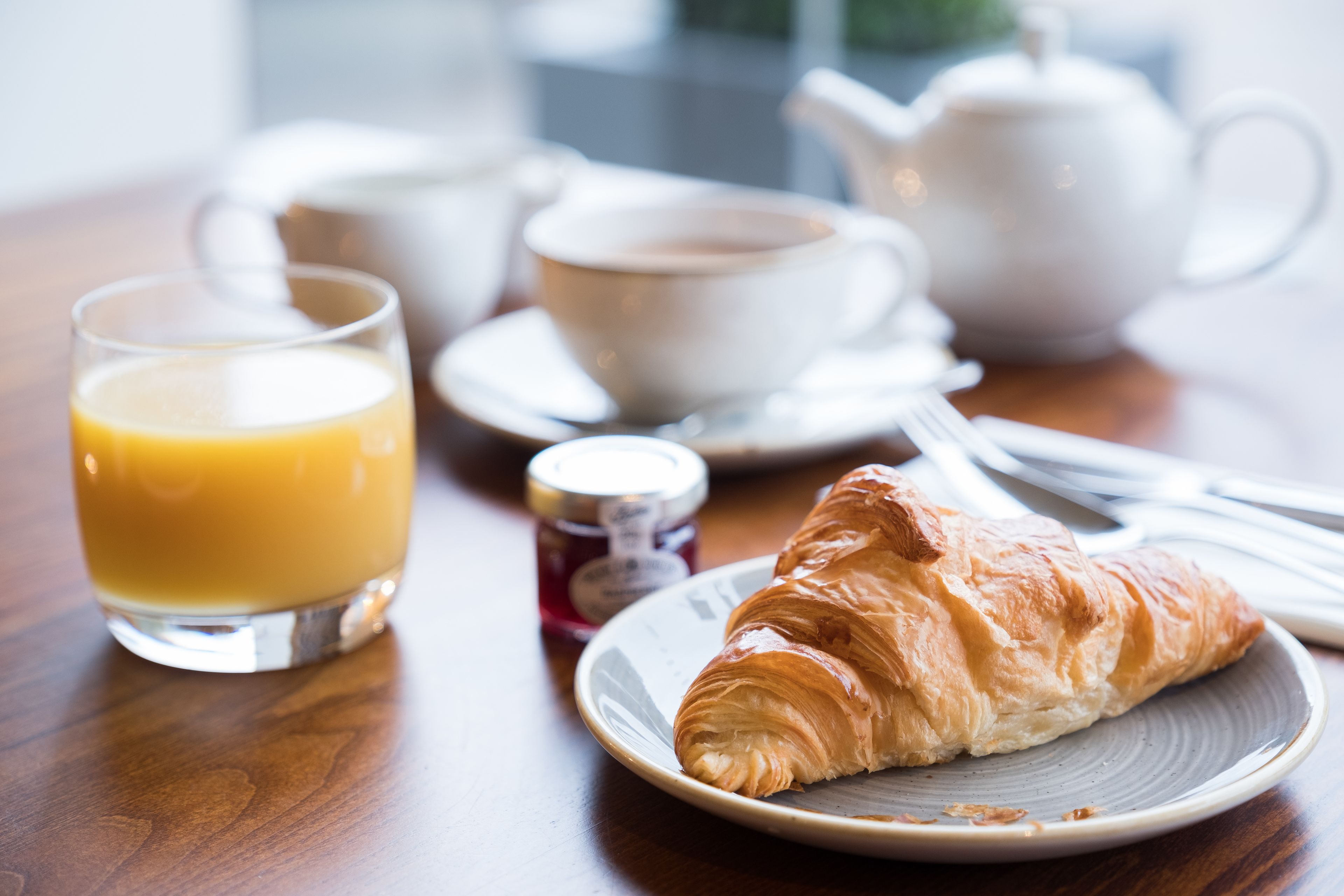 A breakfast setting with a croissant, a glass of orange juice, and a teapot on a wooden table.