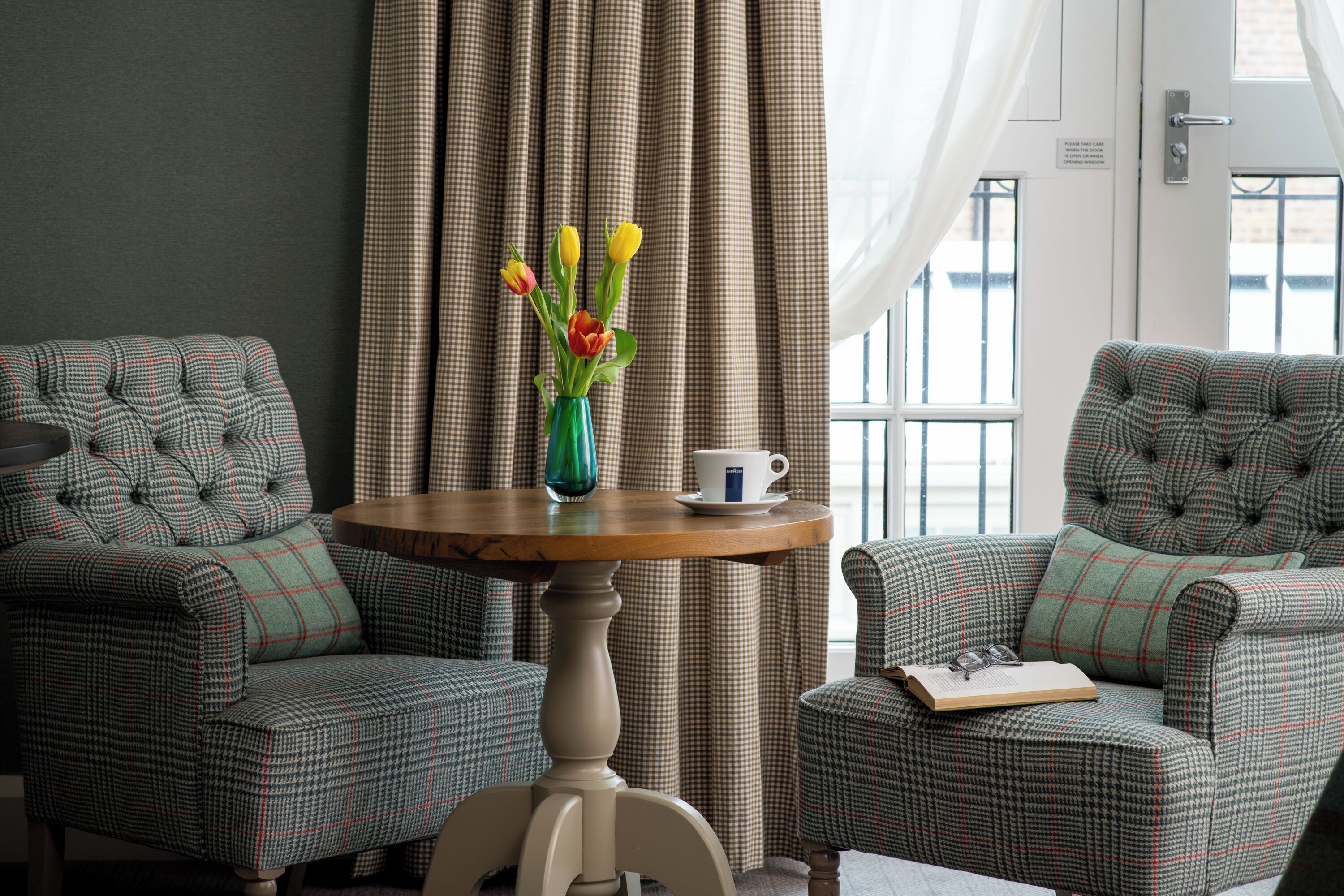 A room with two upholstered armchairs, a round wooden table with a vase of flowers, a cup, and a book.